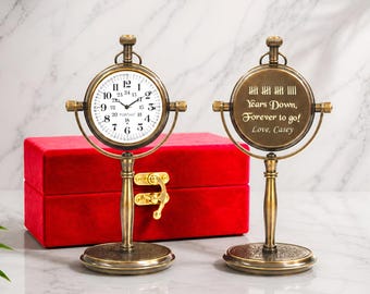 May include: Two antique-style brass desk clocks on stands. One clock face shows numbers 1-24 and the text PORTHO. The other side features engraved tally marks and the text Years Down, Forever to go! Love, Casey. A red velvet box sits behind them.
