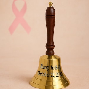 Könnte beinhalten: Eine Messingglocke mit einem dunklen Holzgriff und einer goldfarbenen Oberseite. Auf der Glocke steht "Rang the Bell October 29, 2023". Im Hintergrund befindet sich ein rosa Band.
