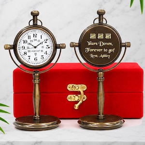 May include: Two antique-style brass desk clocks on stands, one with a white face showing numbers 1-24 and the word PORTHO, the other with an engraved back reading Years Down, Forever to go! Love, Ashley, positioned before a red velvet box.