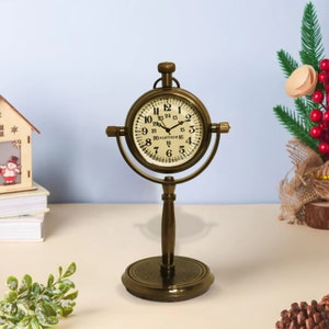K&ouml;nnte beinhalten: Eine goldfarbene Tischuhr mit einem runden Zifferblatt und einem St&auml;nder. Das Zifferblatt der Uhr hat schwarze r&ouml;mische Ziffern f&uuml;r die Stunden und schwarze Markierungen f&uuml;r die Minuten. Das Zifferblatt der Uhr tr&auml;gt auch den Schriftzug "PORTHOS".