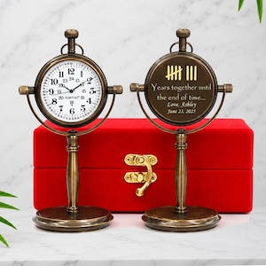 May include: Two antique-style brass desk clocks with circular stands. The left clock features a white face with black numerals and the text PORTHO. The right clock displays tally marks, the text Years together until the end of time... Love, Ashley June 23, 2025.