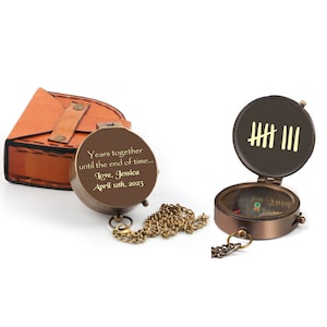 May include: A brass compass with a leather case. The compass face has a traditional design with a compass needle and markings. The lid of the compass has a personalized inscription that reads "Years together until the end of time... Love, Jessica April 12th, 2023". The lid also has five tally marks.