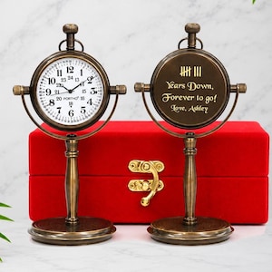 May include: Two antique-style brass desk clocks on stands. One clock features a white face with black numerals and the text PORTHO. The other clock displays the engraved text: IIII Years Down, Forever to go! Love, Ashley. Both clocks are mounted on pedestals.