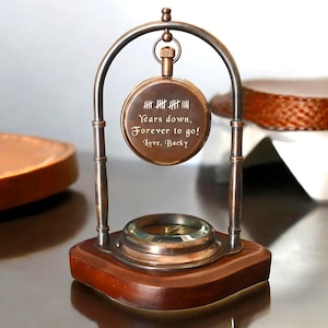 Könnte beinhalten: Eine Tischuhr im Vintage-Stil mit einem Kompass an der Basis und einem Taschenuhr-Zifferblatt, das darüber aufgehängt ist. Das Zifferblatt trägt die Inschrift "Years down, Forever to go! Love, Backy". Die Basis ist aus dunklem Holz.