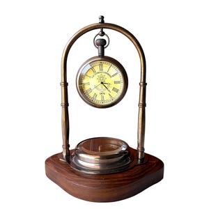 May include: A vintage desk clock with a brass frame and a wooden base. The clock face is cream-coloured with Roman numerals and the text "VICTORIA LONDON 1876". The clock has a pocket watch design and a small ashtray.