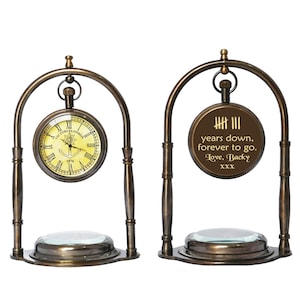 May include: Two antique-style brass clock stands with a compass base. One clock face features Roman numerals and an anchor, the other has an inscription that reads "圭三 years down, forever to go. Love, Backy XXX".