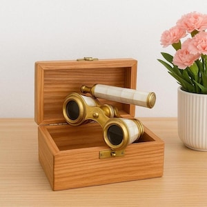 May include: An open wooden box displaying a pair of antique brass opera glasses with white and gold accents. The glasses rest inside the box, which has a brass clasp. A matching handle is also present. The box is made of light-colored wood.