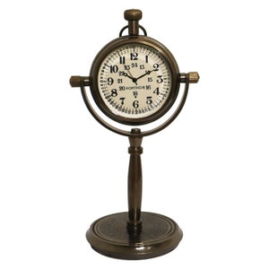 K&ouml;nnte beinhalten: Eine Messing-Tischuhr mit einem runden Zifferblatt und einem St&auml;nder. Das Zifferblatt der Uhr hat schwarze Zahlen und einen wei&szlig;en Hintergrund. Der St&auml;nder ist eine runde Basis mit einer hohen, zylindrischen S&auml;ule.