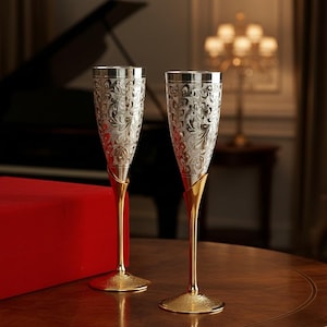 May include: Two champagne flutes with ornate silver-colored bowls and gold-colored stems and bases. The flutes are decorated with a detailed floral pattern. The glasses are displayed on a wooden table.