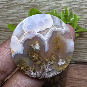 May include: A round, polished agate slice with a unique pattern of white, brown, and gray swirls and dendrites. The agate has a translucent quality, allowing light to pass through it.