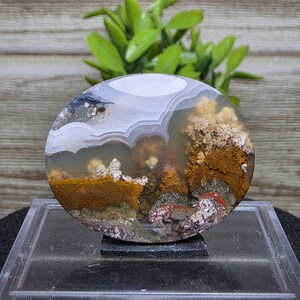 May include: A polished, round agate stone with a landscape-like pattern. The stone features layers of white, brown, orange, and green hues, resembling a miniature natural scene. The agate is displayed on a clear, rectangular stand.