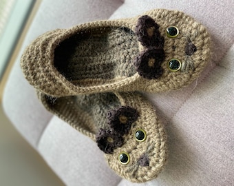 May include: A pair of brown crocheted slippers with a cat face design. The slippers have black felt eyes and a brown felt nose.