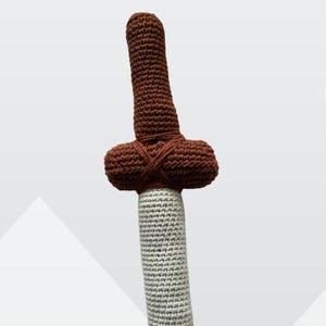 Könnte beinhalten: Ein gehäkeltes Schwertspielzeug mit braunem Griff und Parierstange sowie einer hellgrauen Klinge. Der Griff ist lang und zylindrisch, während die Parierstange horizontal ausgerichtet ist und Garn um die Mitte gewickelt ist, wo sie auf die Klinge trifft.