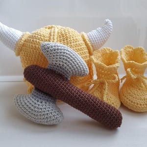 Puede incluir: Un casco de vikingo tejido a ganchillo en color amarillo con cuernos blancos, un par de patucos amarillos a juego con cierre de cordones y un hacha de doble hoja tejida con mango marrón oscuro y hojas gris claro.