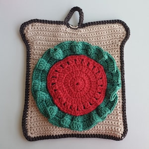 Puede incluir: Un agarrador de olla de ganchillo con forma de rodaja de sandía. La sandía es roja con una cáscara verde y está rodeada por un borde de ganchillo marrón.