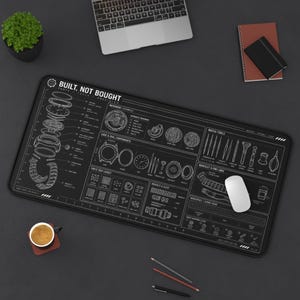 Può includere: Un tappetino da scrivania rettangolare nero con un diagramma tecnico bianco di componenti di orologi, strumenti e istruzioni di montaggio. Il testo "BUILT, NOT BOUGHT" è stampato in alto. Il tappetino include illustrazioni dettagliate di parti.