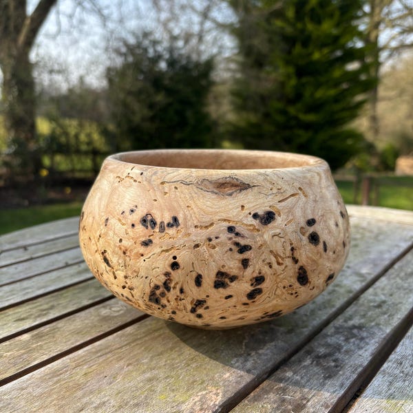 May include: A wooden bowl with a unique burl pattern. The bowl is light brown with dark brown spots and has a rounded shape.