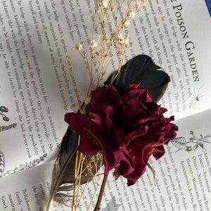 May include: A close-up of a dried red rose with a black feather and white baby's breath flowers on top of an open book. The book is titled "The Poison Garden" and has text about hedge witches and their plant allies.