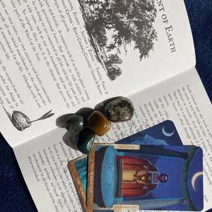 May include: A book open to a page about the element of earth in astrology. The page features text about the element of earth and its connection to the planet Saturn. The book is lying open on a surface with four crystals and two tarot cards, one of which is the Magician card.