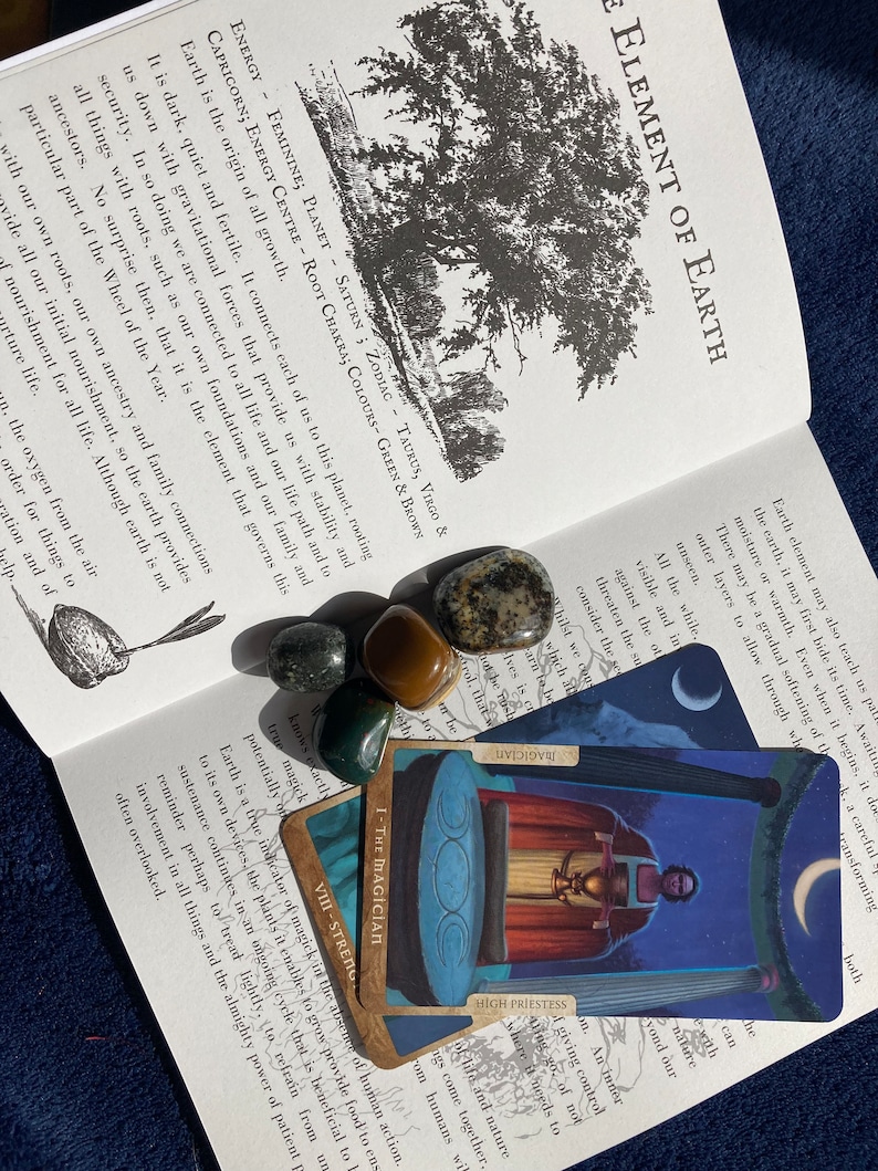 May include: A book open to a page about the element of earth in astrology. The page features text about the element of earth and its connection to the planet Saturn. The book is lying open on a surface with four crystals and two tarot cards, one of which is the Magician card.