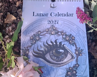 Puede incluir: Un calendario de pared 2027 Lunar Calendar con una ilustración central de un ojo estilizado rodeado por una corona floral. El texto dice The Library of Witchcraft, Lunar Calendar y 2027. El calendario está encuadernado con espiral en la parte superior.