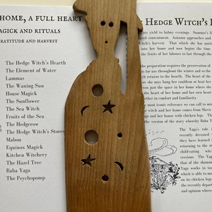 May include: A wooden cutout of a witch's house with a crescent moon, star, and sun cutouts. The text "Hedge Witch's Hearth" is printed on the page next to the cutout.