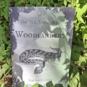 May include: A book cover for "The Witch's Familiars: Woodlanders" featuring a snake illustration. The title and author, Rachael Coats, are in dark grey on a light green background. The book is surrounded by green foliage.