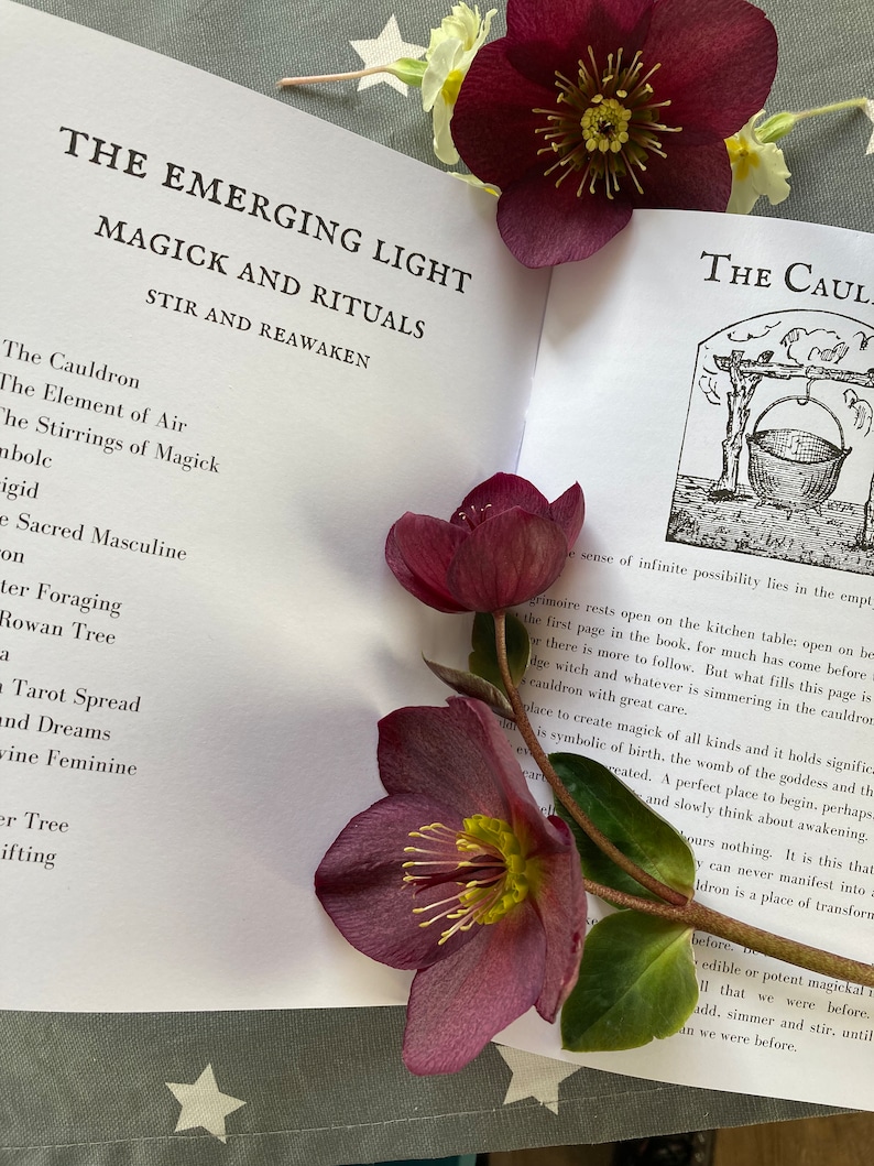 May include: An open book with the title "The Emerging Light Magick and Rituals Stir and Reawaken" and a section titled "The Cauldron". The book is lying on a surface with a few purple and white flowers.