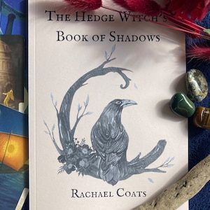 May include: A book titled "The Hedge Witch's Book of Shadows" by Rachael Coats. The cover features a black raven perched on a branch with flowers and leaves. The background is a light beige color.