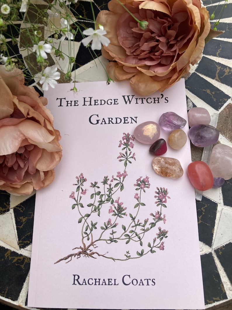 May include: A pink book with the title "The Hedge Witch's Garden" by Rachael Coats. The book is surrounded by pink and white flowers and various crystals.