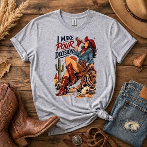 Könnte beinhalten: Graues T-Shirt mit dem Text "I Make Pour Decisions" und einer Grafik einer Cowgirl, die aus einer Flasche trinkt. Die Cowgirl trägt einen roten Hut, ein blaues Hemd und braune Stiefel. Ein Kaktus und eine Wüstenlandschaft sind im Hintergrund.