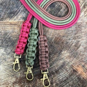 May include: Three lanyards crafted from woven paracord in magenta, olive green, and brown. Each lanyard features a braided section and a brass-toned metal swivel lobster clasp at the end. The cords are coiled together in a circular arrangement.