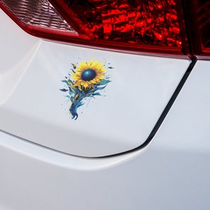 K&ouml;nnte beinhalten: Ein wei&szlig;es Auto mit einem blauen und gelben Sonnenblumen-Aufkleber auf der hinteren Sto&szlig;stange.