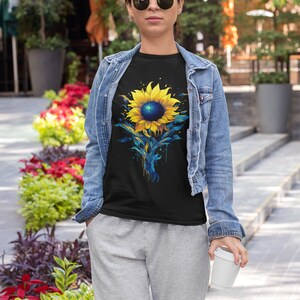 K&ouml;nnte beinhalten: Ein schwarzes T-Shirt mit einem bunten Sonnenblumen-Motiv. Die Sonnenblume ist in Gelb-, Blau- und Gr&uuml;nt&ouml;nen bemalt. Das Shirt wird von einer Person getragen, die eine Jeansjacke und graue Jogginghosen tr&auml;gt.