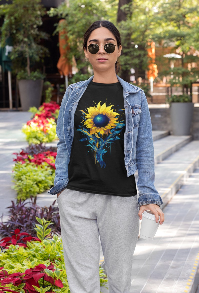 K&ouml;nnte beinhalten: Ein schwarzes T-Shirt mit einem bunten Sonnenblumen-Motiv. Die Sonnenblume ist in Gelb-, Blau- und Gr&uuml;nt&ouml;nen bemalt. Das Shirt wird von einer Person getragen, die eine Jeansjacke und graue Jogginghosen tr&auml;gt.