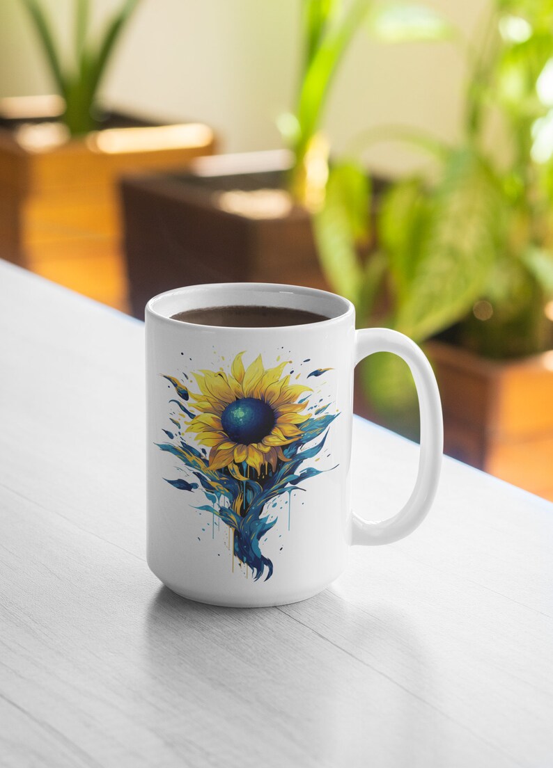 K&ouml;nnte beinhalten: Wei&szlig;e Keramiktasse mit einem blauen und gelben Sonnenblumen-Design. Die Sonnenblume hat eine dunkelblaue Mitte und gelbe Bl&uuml;tenbl&auml;tter. Die Tasse ist mit Kaffee gef&uuml;llt.
