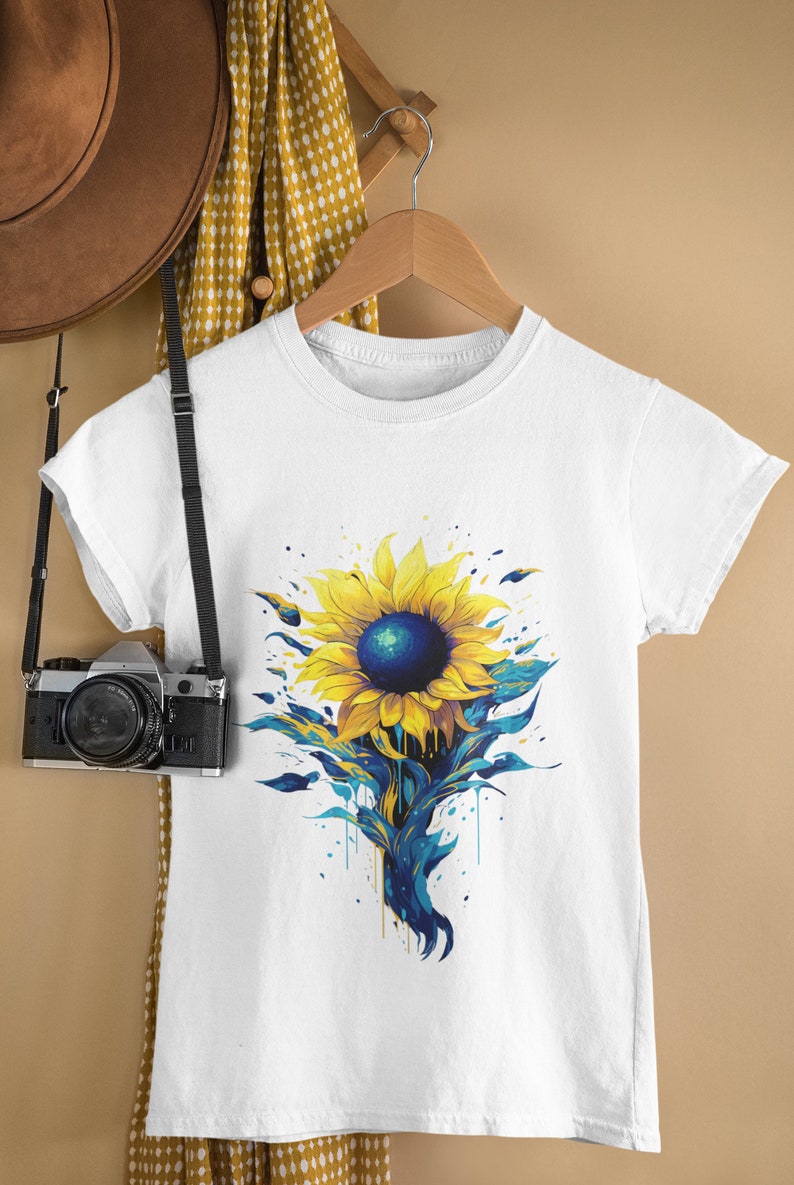 K&ouml;nnte beinhalten: Wei&szlig;es T-Shirt mit einem blauen und gelben Sonnenblumen-Motiv. Die Sonnenblume hat eine blaue Mitte und gelbe Bl&uuml;tenbl&auml;tter mit blauen Farbstichen.