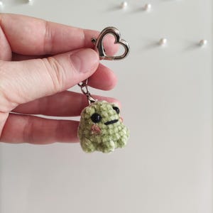 May include: Green plush frog keychain with a heart-shaped metal ring. The frog has black eyes and a small smile.