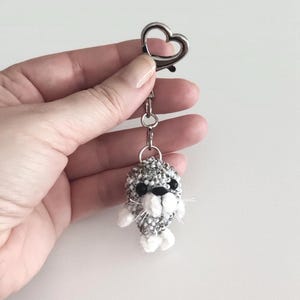 May include: A handmade keychain featuring a beaded seal charm. The seal has a gray and white beaded body, black eyes, and white accents. The keychain includes a silver-tone heart-shaped clasp and a chain.