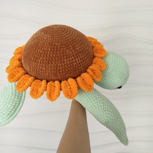 May include: A crocheted turtle toy with a brown shell and a green body. The shell is decorated with a bright orange sunflower.