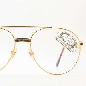 May include: Gold-rimmed aviator eyeglasses with clear lenses. The frames have a double bridge and brown temple tips. A sticker on the lens reads "Vintage eyewear Collection".