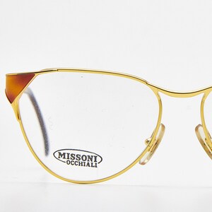 May include: Gold-tone metal spectacles with a thin frame and cat-eye silhouette. The temples feature brown accents at the outer corners. The right lens displays the text MISSONI OCCHIALI in black lettering. These glasses have clear lenses.