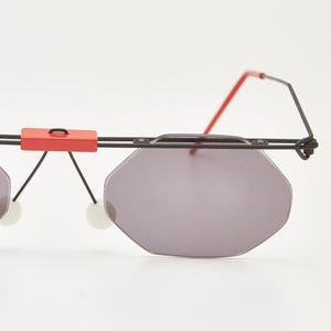 Puede incluir: Gafas de sol geométricas con montura de alambre negro y detalles rojos. Las lentes son de color gris apagado y tienen forma octogonal. El puente de las gafas tiene un detalle rectangular rojo.