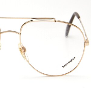 Pode incluir: Óculos de aviador dourados com lentes transparentes. Os óculos têm uma ponte dupla e uma armação fina. O nome da marca "MENRAD" está impresso na lente inferior direita. As hastes são marrons escuros.