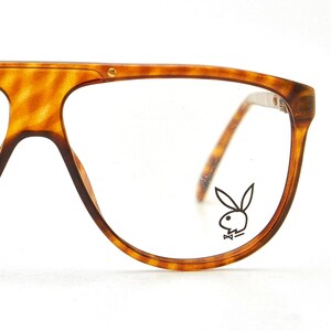 May include: Close-up of vintage eyeglasses with a tortoise shell pattern. The frames are thick and rectangular, with a small gold accent on the top corner. The right lens features the Playboy bunny logo.