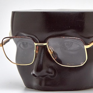 May include: Gold-rimmed eyeglasses with a square shape are displayed on a dark brown head-shaped display. The glasses have a classic design with a gold frame and tinted lenses. The display is smooth and has facial features.