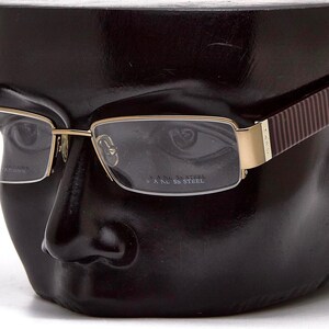 Könnte beinhalten: Goldgerahmte Brille mit rechteckigen Gläsern. Die Bügel sind braun mit vertikalen Streifen. Die Brille wird auf einem schwarzen Schaufensterkopf präsentiert. Der Text "5 A NL SS STEEL" ist auf dem Glas zu sehen.
