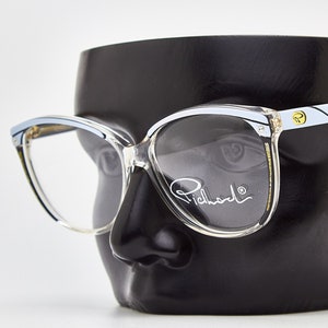 May include: Clear plastic cat-eye glasses with a white and blue trim. The glasses have a gold logo on the right temple and the word "Richardo" written on the right lens.