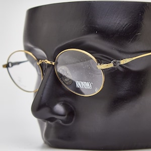 Op de afbeelding: Ronde bril met gouden rand en zwarte pootjes. Het woord "Anonimo" staat op de lens gedrukt.