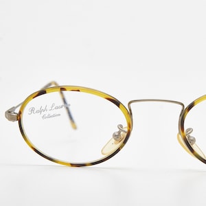 Puede incluir: Gafas de forma ovalada con un patrón de carey en las monturas y varillas. La montura metálica es dorada y las lentes son transparentes. El texto "Ralph Lauren Collection" es visible en la lente izquierda.